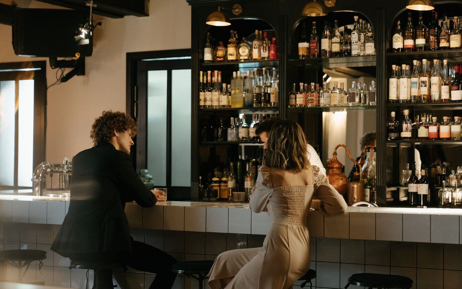 Two people sit at a bar counter lined with shelves of liquor bottles
