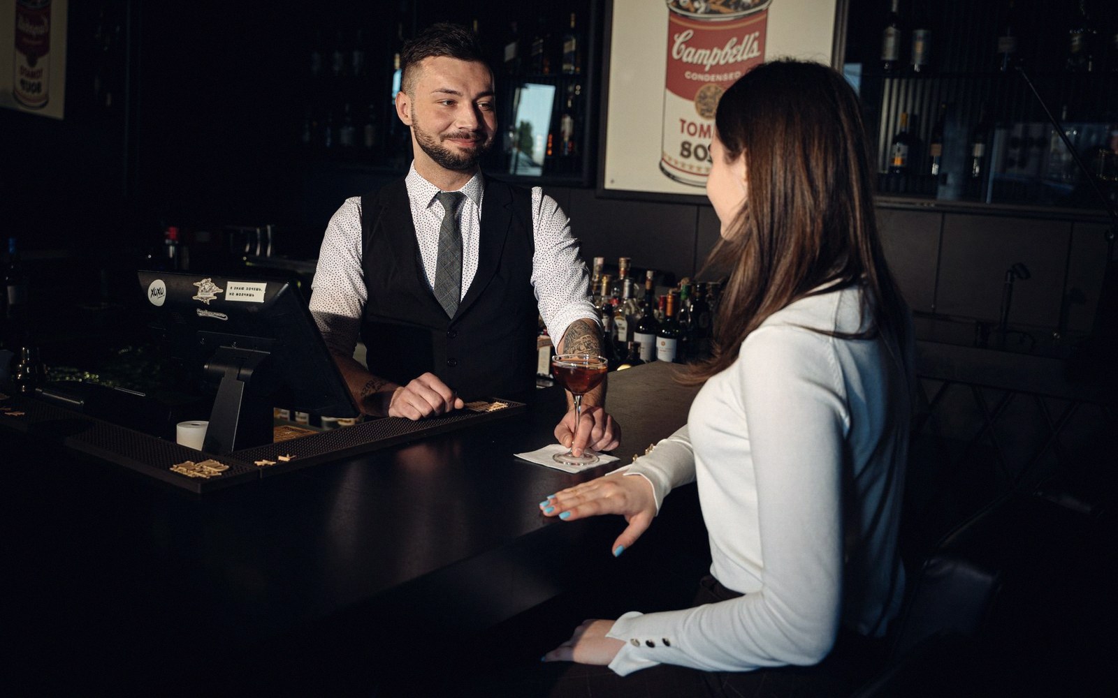 Bartender in a vest and tie serves a cocktail to a seated customer at the bar