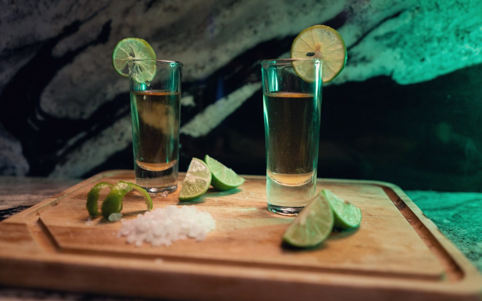 Two tequila shots with lime wheels on rims, salt and lime wedges on a wooden board