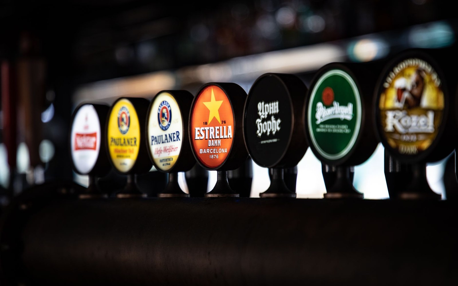 A row of beer tap handles including Paulaner, Estrella Damm and Kozel at a bar