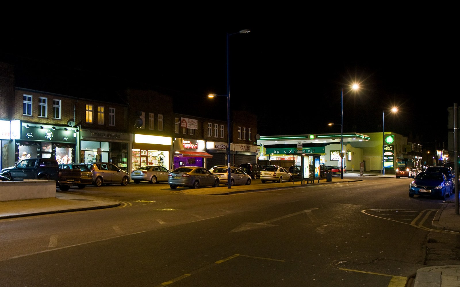 Parked cars line a quiet high street at night beside a lit bp petrol station