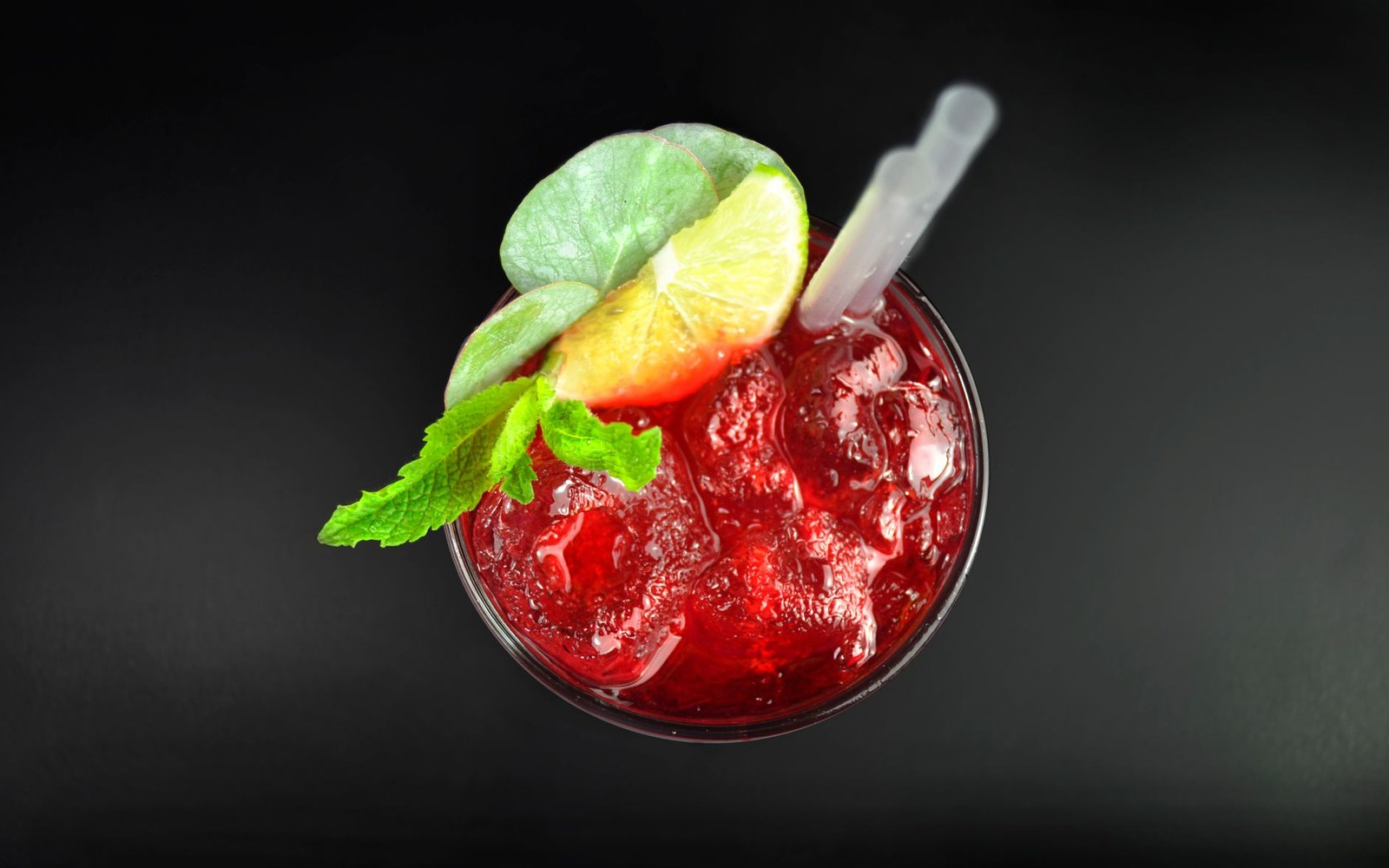 Overhead view of a red cocktail over ice, garnished with lime wedge, mint, and straw