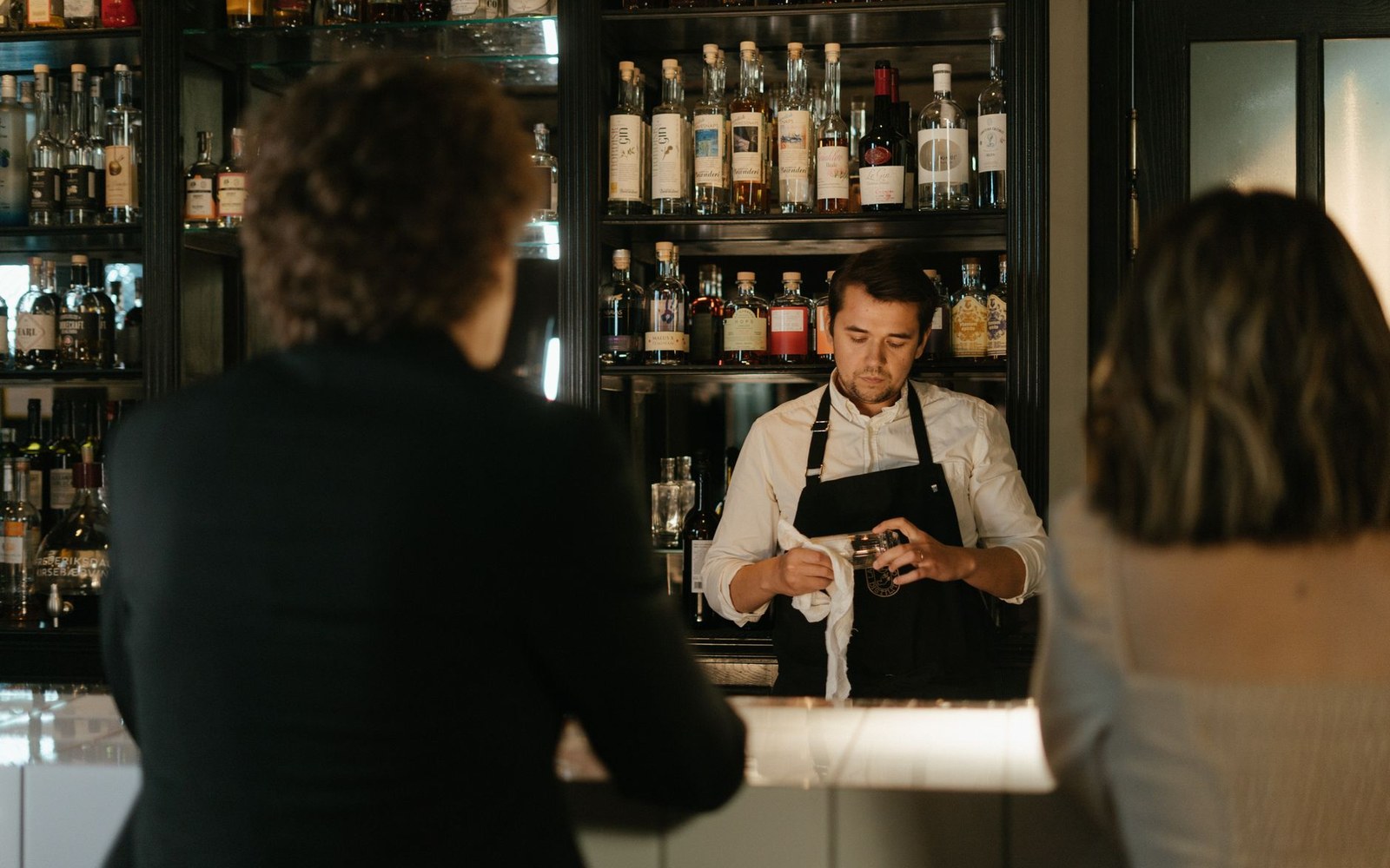 Bartender in an apron polishes a glass behind shelves of bottles