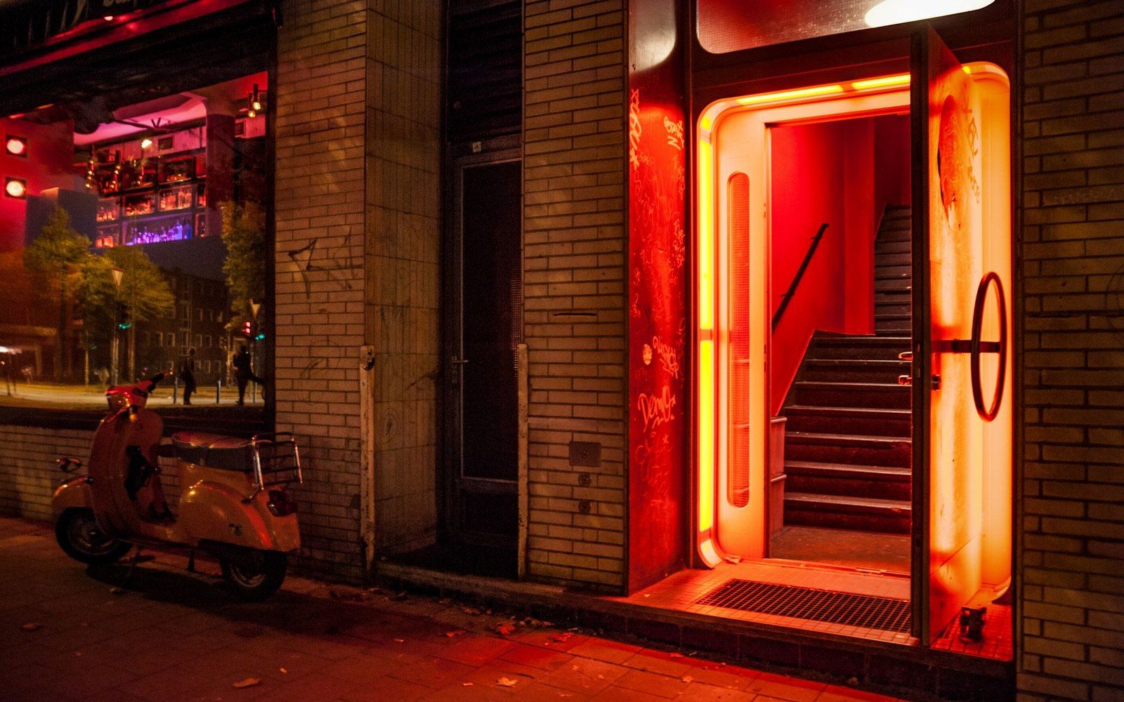Neon-lit doorway with staircase glows red beside a parked scooter at night