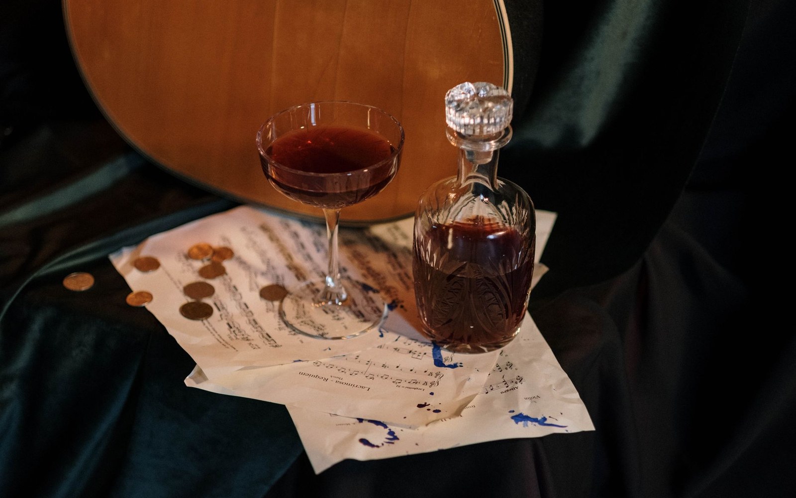 A dark cocktail in a coupe glass beside a decanter, coins, and sheet music