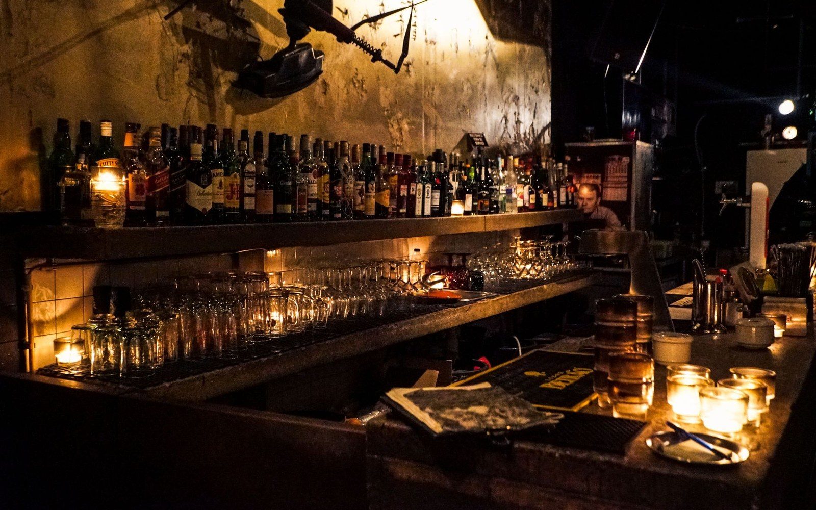 Bottles line a shelf above a candlelit bar with stacked glasses and a bartender
