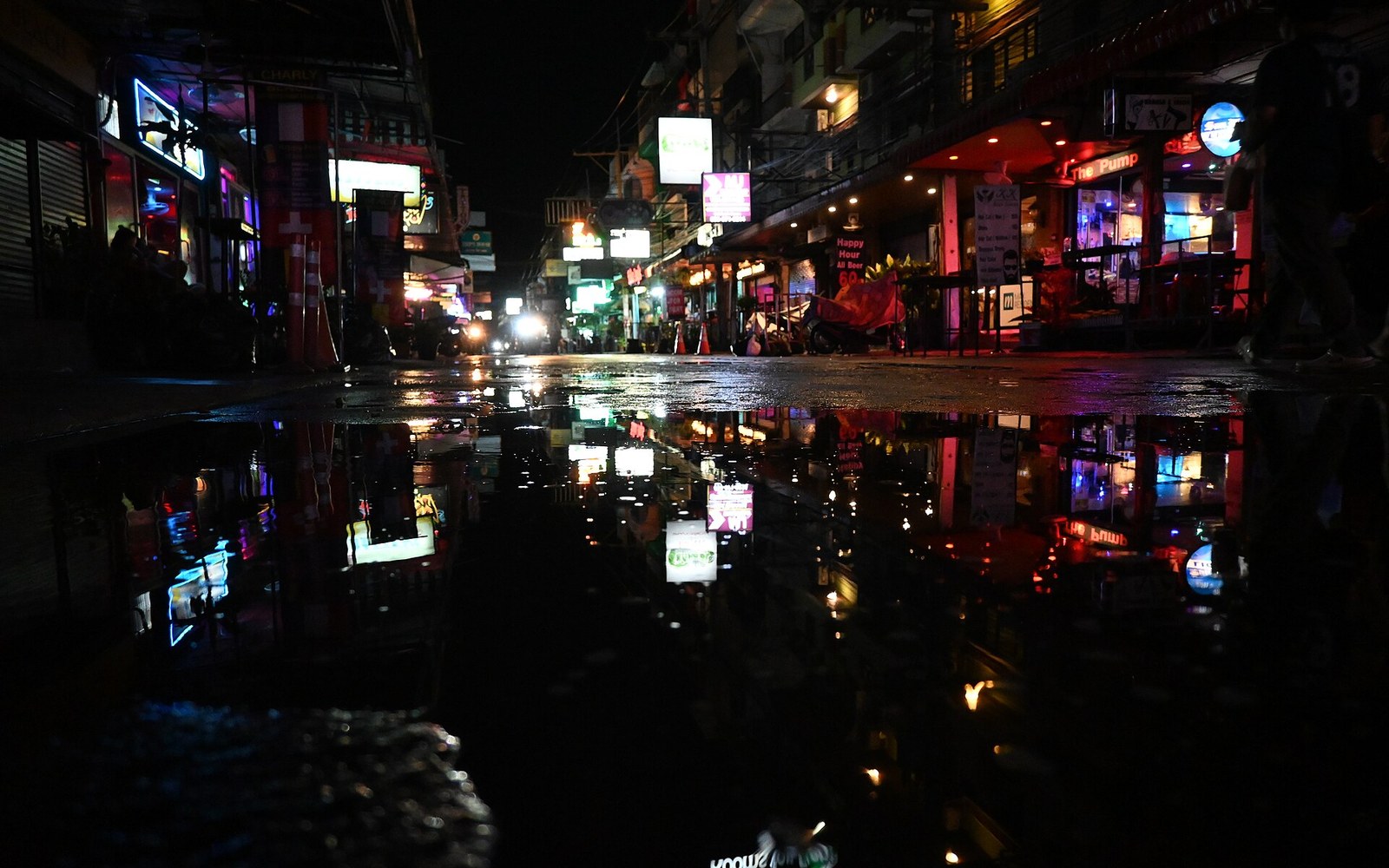 A bar's neon sign reflected in wet pavement outside, shot from low angle, rain still falling
