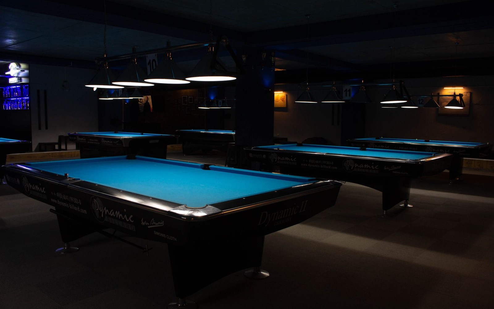 A pool table under a single hanging light, the rest of the room in shadow, one cue resting against the rail